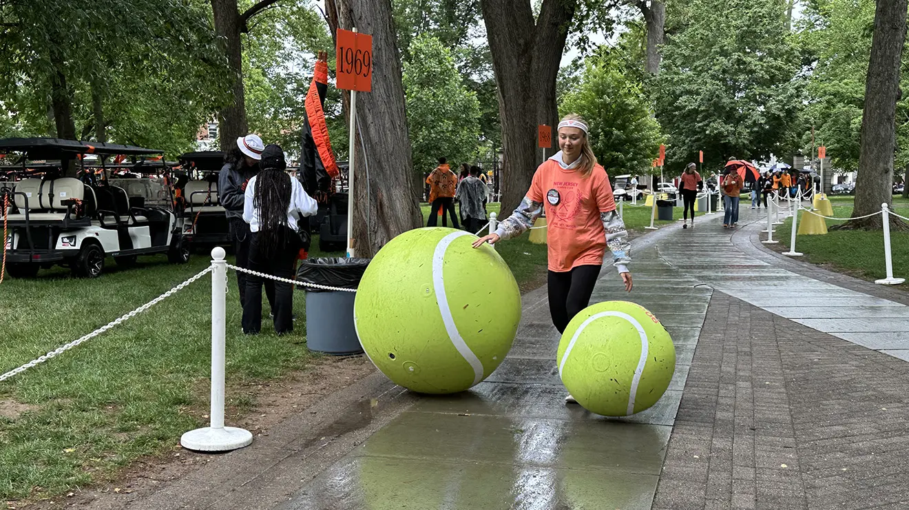 JulieReunions4_cropped.jpg A woman stands between two giant tennis balls, one coming up to her knee and the other above her hip.